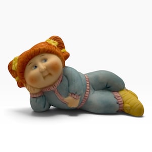 Könnte beinhalten: Eine Keramikfigur einer Puppe mit orangefarbenen Zöpfen, die mit gelben Schleifen gebunden sind. Sie trägt ein hellblaues langärmeliges Outfit mit rosa Besatz und gelben Schuhen und liegt in einer ruhenden Position auf der Seite.