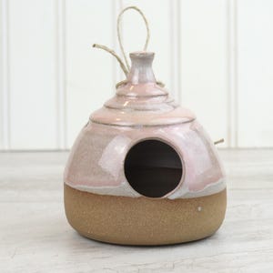 Pode incluir: Um viveiro de cerâmica com uma abertura redonda e uma argola para pendurar. O viveiro é principalmente rosa com uma base marrom e uma argola de barbante natural. O viveiro foi projetado para atrair pássaros.