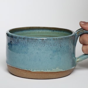 May include: A wide, low-profile ceramic mug featuring a glossy, variegated blue glaze that transitions into a natural, unglazed tan clay base. The mug has a sturdy, integrated handle and a smooth, rounded rim, showcasing a handcrafted aesthetic.