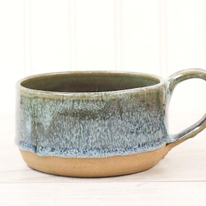 May include: A wide ceramic mug featuring a reactive glaze with shades of deep green and blue that transition into a textured, speckled finish. The lower portion of the mug remains unglazed, revealing a natural, matte tan clay base.