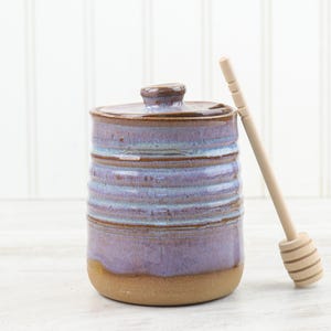 Könnte beinhalten: Keramik-Honigtopf mit Deckel, lavendel- und braun glasiert. Ein Honiglöffel aus Holz liegt neben dem Topf. Der Topf hat eine zylindrische Form mit horizontalen Rillen. Der Boden ist naturfarben, unglasiert.
