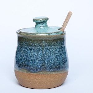 May include: A ceramic honey pot with a lid and a wooden dipper. The pot features a textured blue and teal glaze on the upper portion and a natural brown clay base. The lid is a matching teal color with a small knob handle.