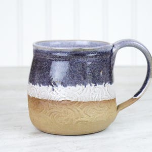 May include: A handmade ceramic mug with a purple, white, and brown glaze. The mug has a textured design and a rounded shape.