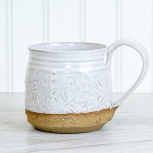 May include: White ceramic mug with a textured floral pattern on the upper portion and a tan base. The mug has a curved handle and is suitable for hot or cold beverages. The mug is handmade and has a rustic, artisanal appearance.