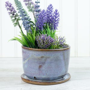 Puede incluir: Una maceta de cerámica con un esmalte lavanda, que contiene lavanda artificial y follaje verde. La maceta se asienta sobre un platillo a juego. La maceta tiene un borde y una base marrones.