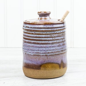 May include: A handmade ceramic jar with a lid and a wooden spoon. The jar has a cylindrical shape with horizontal lines and a colour gradient of brown, purple, and tan. The spoon is inserted into a hole in the lid.