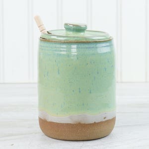 May include: A ceramic jar with a lid and a small wooden spoon. The jar is cylindrical with a light green glaze, a white band, and a brown base. The spoon rests on the lid.