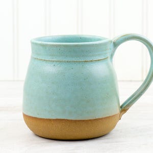 May include: A handmade ceramic mug with a rounded base and a large handle. The mug features a light blue glaze on the upper portion and a natural tan color on the lower portion. The mug is approximately 4 inches tall.