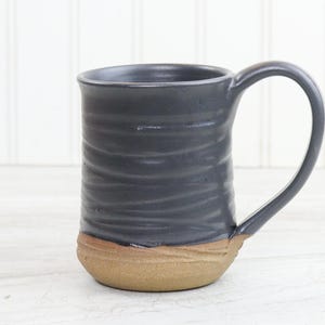May include: A handmade ceramic mug with a dark gray upper and a tan base. The mug has a curved handle and a textured, ridged surface. The mug is approximately 5 inches tall.