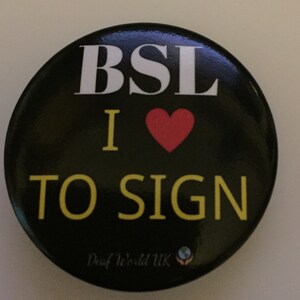 Può includere: Bottone nero con testo bianco che dice "BSL I ♥ TO SIGN" e il testo "Deaf World UK" con un'icona del globo terrestre sotto.