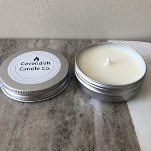May include: A silver tin candle with a white label that reads "Cavendish Candle Co." and a flame icon. The candle is in a silver tin with a white wax top and a white wick.