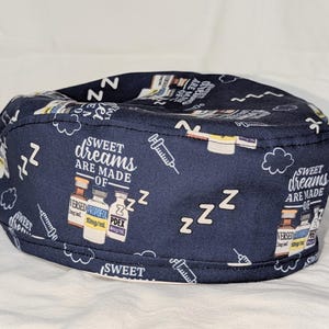 May include: Navy blue scrub cap with a repeating pattern of syringes, clouds, and the text "Sweet Dreams Are Made Of" with images of medication vials. The cap also features the letter "Z" repeated throughout.