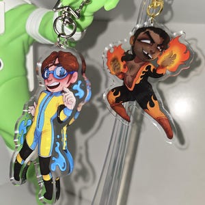 May include: Two acrylic keychains featuring illustrated characters. One character wears a yellow and blue suit with blue goggles, surrounded by blue liquid. The other character wears a black and orange flame-patterned suit with fire effects.