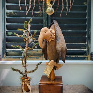 May include: A brown fabric bird with a long beak and a ruffled collar stands on a wooden base. The bird is holding a sign that says "SMILE PUZZLES". The bird is in front of a window with a garland of dried gourds hanging from the top.