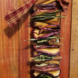 May include: A rustic, homemade garland made with dried apple slices, cinnamon sticks, and bay leaves. The garland is tied with a red and brown plaid ribbon and is hanging on a wooden door.
