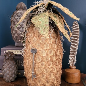 May include: A brown, textured pineapple-shaped object with a key sticking out of it. The object is sitting on a wooden table with a book and a feather quill in the background.