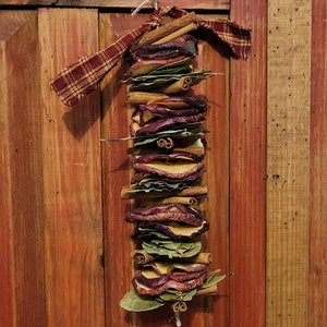 May include: A rustic, homemade ornament made with dried apple slices, cinnamon sticks, and bay leaves. The ornament is tied with a red and black plaid ribbon and hangs from a string.
