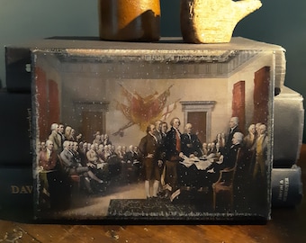 May include: A framed painting depicting a group of men in historical clothing gathered around a table signing a document. The painting is set in a room with a large flag hanging on the wall. The painting is framed in a dark wood frame.