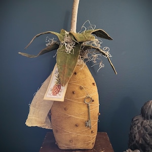 May include: A handmade fabric pineapple with a key and a tag that says "Pineapple" in brown lettering. The pineapple is decorated with brown twine and brown cloves. The pineapple is sitting on a wooden surface.