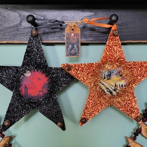May include: Two glittery star-shaped ornaments with Halloween themes. One is black with a red and black scene of a house and a bat, the other is orange with a scene of a house and people walking down a path.