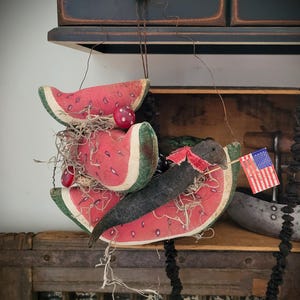 May include: A whimsical watermelon-themed decoration featuring three slices of watermelon with red and green details. A black bird-like figure holds a tiny American flag. Decorative berries and dried moss add texture.