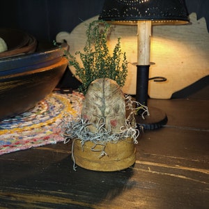 May include: A small, rustic wooden box with a fabric top featuring a tree design and red accents. The box is filled with dried moss and topped with a sprig of green foliage.