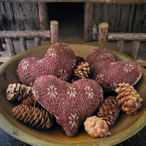 May include: A wooden bowl filled with heart-shaped fabric ornaments and pine cones. The ornaments are burgundy with a white snowflake pattern. The bowl is light brown, and the pine cones are various shades of brown.