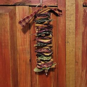 May include: A rustic, homemade ornament made with dried apple slices, cinnamon sticks, and bay leaves. The ornament is tied with a plaid fabric ribbon and hangs from a wooden surface.