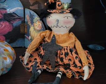 May include: A fabric doll featuring a pointed hat with orange and black patterns, a yellow checkered long-sleeve top, and an orange skirt with black cat silhouettes. The doll has a stitched face, a dark star ornament, and black and white striped legs.