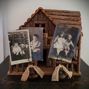 May include: A rustic wooden photo holder shaped like a cabin, displaying three vintage black and white photographs of children. The holder has a natural bark finish and includes metal clips and decorative fabric.