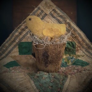 May include: A yellow chick sitting in a brown woven nest, with a brown burlap base. The chick is sitting on a patchwork quilt with green, brown, and white fabric.