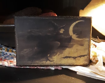 May include: A vintage-style image of a witch flying on a broomstick with a crescent moon in the background. The image is printed on a dark brown wooden plaque.