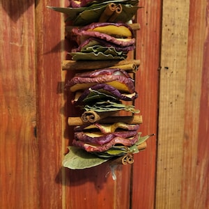 May include: A stack of dried apple slices and cinnamon sticks, layered with green leaves, hanging on a wooden wall.