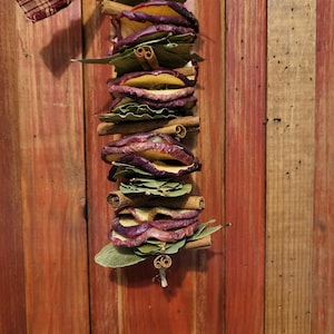 May include: A dried apple and cinnamon stick garland with green leaves. The garland is hanging from a red and white plaid ribbon.