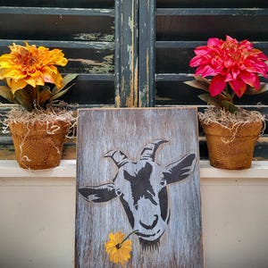 May include: A rectangular wooden sign featuring a black graphic illustration of a goat head with a yellow flower in its mouth, flanked by two potted artificial flowers in burlap-wrapped pots, one with yellow petals and one with bright pink petals.