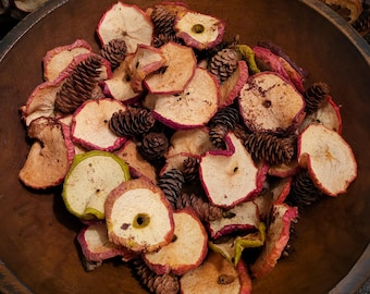 May include: A large, dark brown wooden bowl filled with a collection of dried apple slices and small brown pinecones. The apple slices feature light-colored centers with reddish-pink edges, while the pinecones are textured and varied in size.