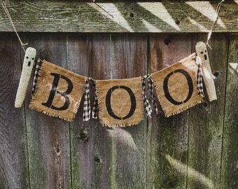 May include: A burlap banner with the word "BOO" spelled out in black letters. The banner is decorated with black and white checkered ribbon and is hanging from a wooden fence.