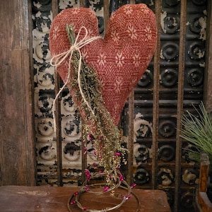 May include: A large, heart-shaped decorative item made of red fabric with a floral pattern. It is adorned with dried greenery and tied with twine. The heart sits on a metal stand with a circular base, decorated with pink beads.