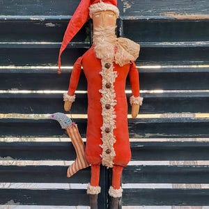 May include: A handmade Santa Claus doll with a long red coat, hat, and beard. The doll has a cream-colored beard and trim, with brown button details. It holds a striped stocking. The doll is a festive holiday decoration.