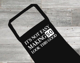 May include: Black apron with the text "IT'S NOT EASY MAKING 50 LOOK THIS GOOD" in white. The apron has a rectangular neck and a long body. The number 50 is in a white box.
