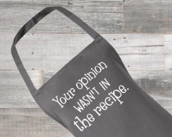 May include: Charcoal gray apron with the text "Your opinion WASN'T IN the recipe" in white script. The apron has a loop for the neck and ties at the waist. The background is a light gray wooden surface.