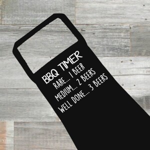 May include: Black apron with white text reading "BBQ TIMER". The text continues with "RARE... 1 BEER", "MEDIUM... 2 BEERS", and "WELL DONE... 3 BEERS". The apron is displayed on a wooden background.