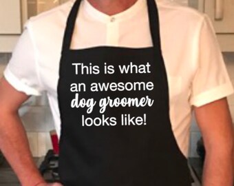 May include: Black apron with the text "This is what an awesome dog groomer looks like!" in white. The apron has a neck strap and waist ties. The person is wearing a white shirt.