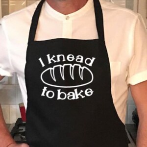 May include: Black apron with the text "I knead to bake" in white, featuring a bread loaf illustration. The apron is worn over a white shirt. A kitchen accessory for baking.