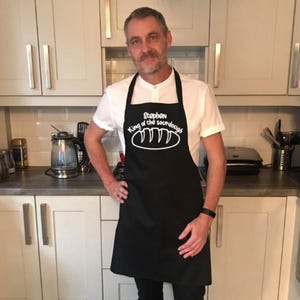 May include: A black apron with white text that reads "Stephen King of the sourdough" and a white illustration of a loaf of bread. The apron is being worn by a person in a white shirt.