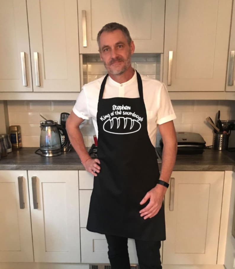 May include: A black apron with white text that reads "Stephen King of the sourdough" and a white illustration of a loaf of bread. The apron is being worn by a person in a white shirt.