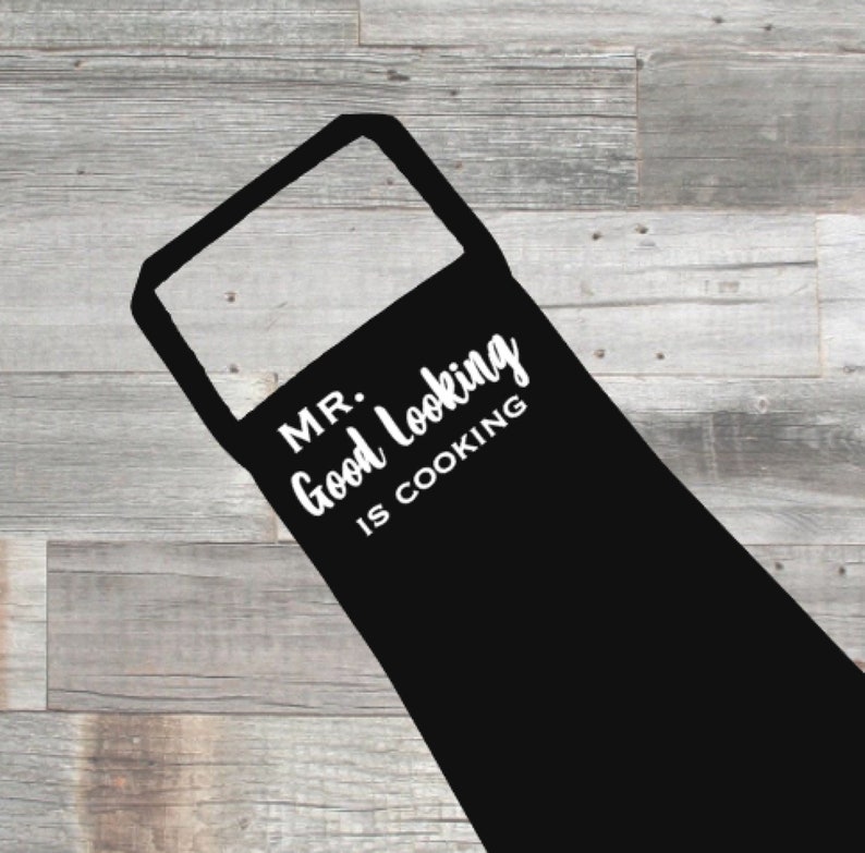 May include: Black apron with the text "MR. Good Looking IS COOKING" in white. The apron has a rectangular bib and a loop for the neck. The background is a light gray wooden surface.