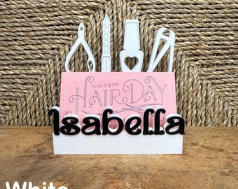 May include: White business card holder with the text "have a great hair day" and the name "Isabella" in black letters. The holder features a design of hairdressing tools, including scissors, a comb, and a nail file.