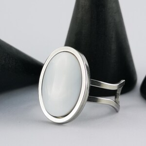 Könnte beinhalten: Ein silberner Ring mit einem ovalen, weißen Stein, der in einen silbernen Rahmen gefasst ist. Der Ring hat ein geteiltes Schaftdesign. Im Hintergrund befinden sich schwarze, kegelförmige Objekte.