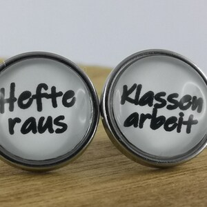 Könnte beinhalten: Zwei silberfarbene Ohrringe mit weißen Glascabochons. Der linke Ohrring sagt "Heft raus" und der rechte Ohrring sagt "Klassenarbeit".